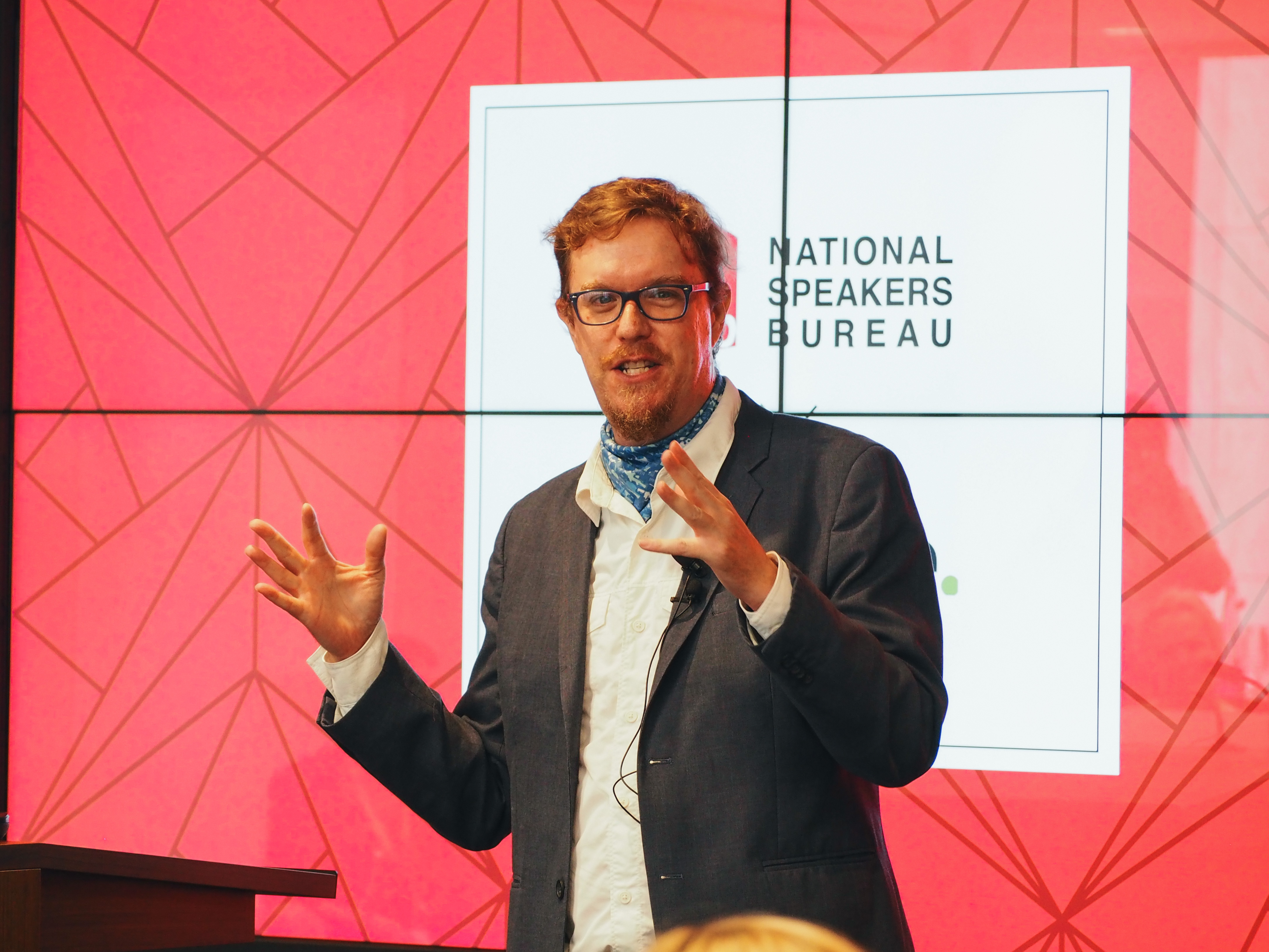 Jesse Hirsh speaking at a podium in front of a National Speakers Bureau screen.