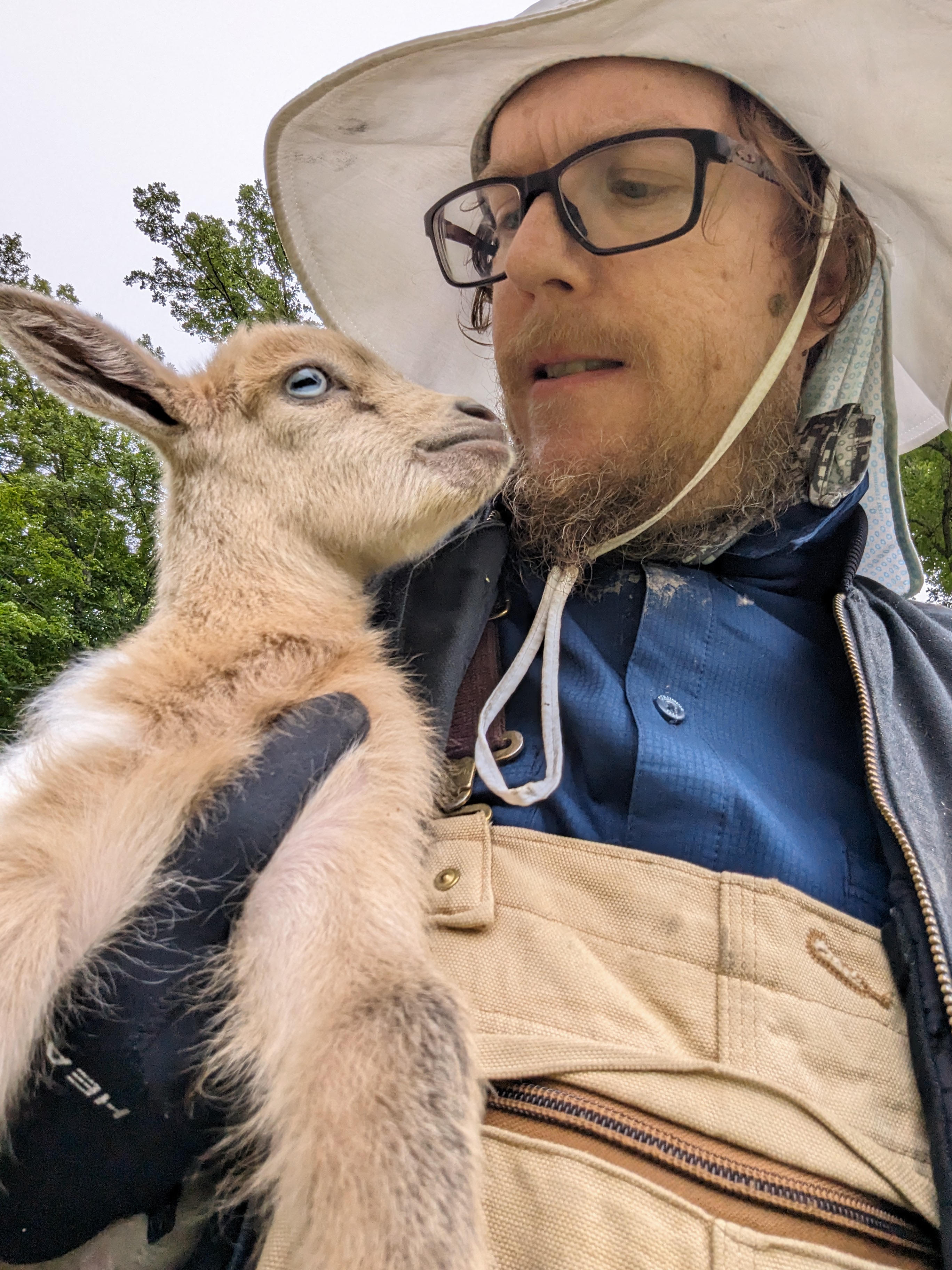 Jesse Hirsh holding a young goat close to his face outdoors.